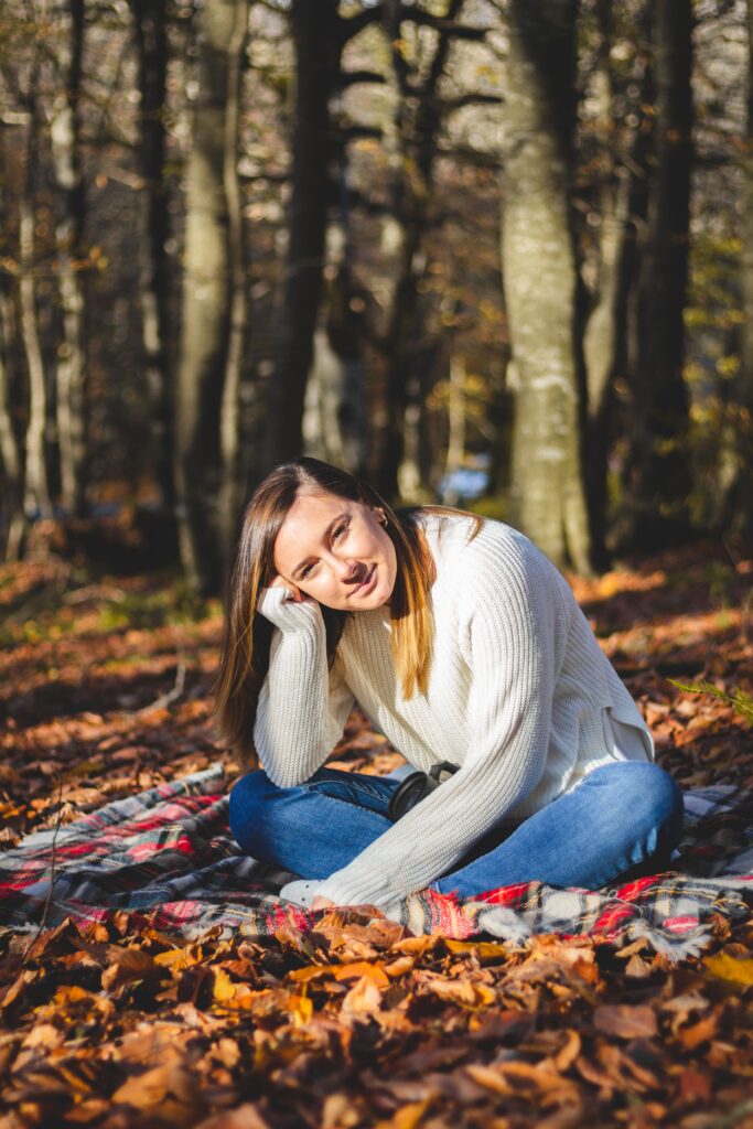 A woman in a cozy sweater sits among fallen leaves in a tranquil autumn forest.